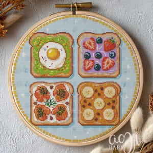 Op de afbeelding: Kruissteekborduurwerk in een houten hoepel met vier sneetjes toast met verschillende toppings. Eén heeft een ei, een ander bessen, een derde tomaten en de laatste bananen. De achtergrond is lichtblauw met een stippenpatroon.