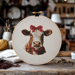 May include: A close-up of a cross-stitch embroidery of a brown and white cow with a red bow on its head. The embroidery is framed in a wooden hoop.