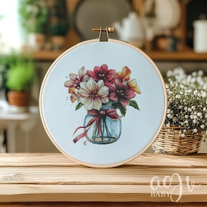 Op de afbeelding: Een borduurring met kruissteek met een witte achtergrond waarop een boeket roze, witte en oranje bloemen in een glazen pot met een bruin lint te zien is.