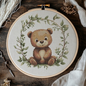 Puede incluir: Un oso de peluche marrón sentado en una corona de hojas verdes. El oso sonríe y tiene una nariz marrón claro y ojos marrones.