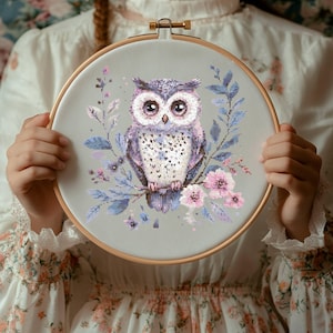 Puede incluir: Un patrón de punto de cruz con un búho morado y blanco con flores rosas y hojas azules sobre un fondo gris. El búho está sentado en una rama con las alas plegadas. El patrón está enmarcado en un aro de madera.