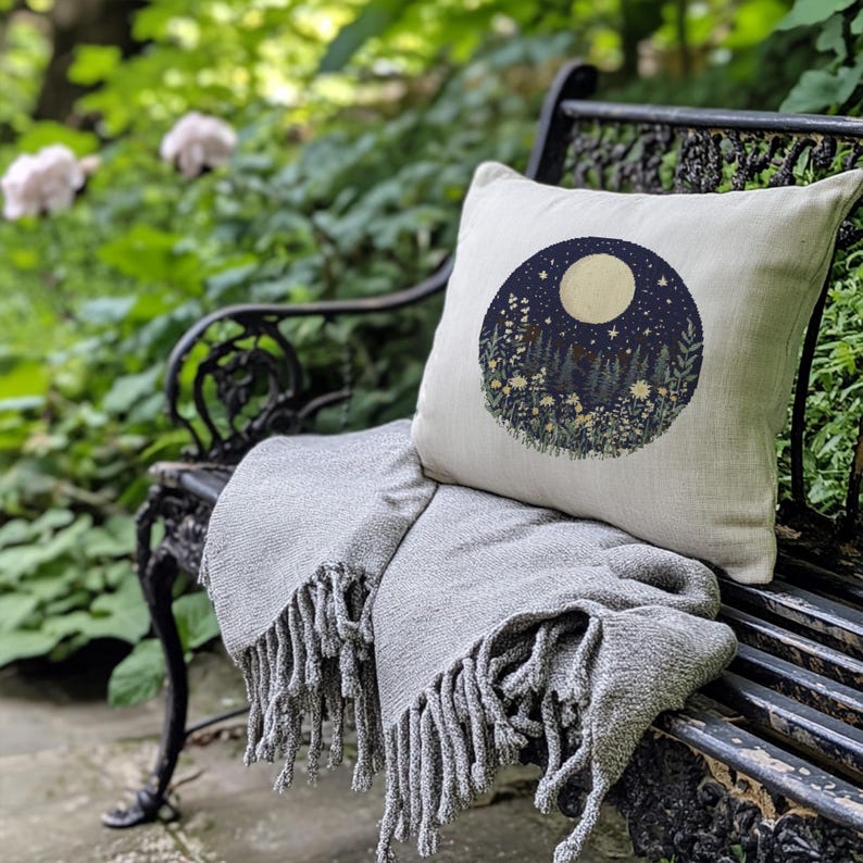 May include: A white throw pillow with a circular design of a moon, stars, and a forest scene in shades of blue, green, and yellow. The pillow is sitting on a black metal bench with a gray throw blanket.