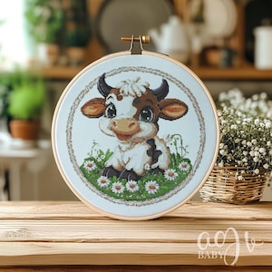 May include: A cross-stitch embroidery hoop with a white background featuring a cartoon cow with brown spots and big eyes. The cow is surrounded by green grass and white daisies.