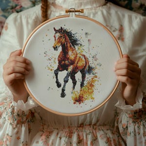 Puede incluir: Una bordado de punto de cruz de un caballo marrón en un aro blanco con un fondo colorido de amarillo, naranja y rosa.