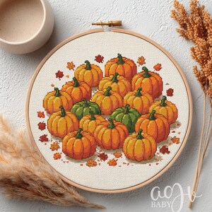 以下が含まれることがあります： オレンジと緑のカボチャのクロスステッチ刺繍フレーム。カボチャは秋の葉に囲まれ、クリーム色の生地にセットされています。刺繍は木製のフレームで囲まれており、秋の装飾に最適で、直径は約20cmです。