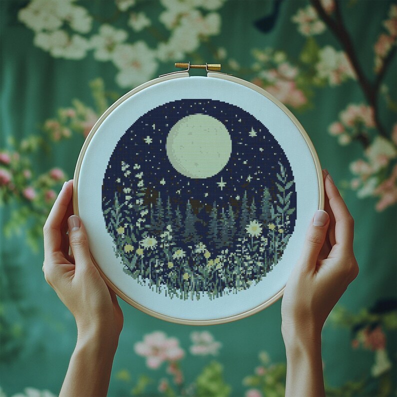 May include: A cross-stitch embroidery hoop with a design of a full moon, stars, and a forest scene with flowers. The moon is white, the stars are yellow, and the forest is green and blue.