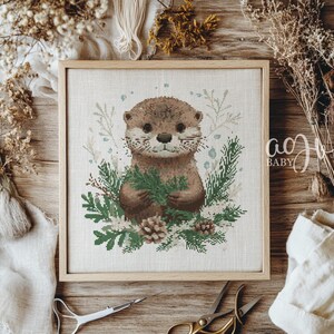 May include: A cross-stitch pattern featuring a brown and white otter holding a sprig of green leaves. The otter is surrounded by a wreath of green leaves and white flowers. The pattern is framed in a light brown wood frame.