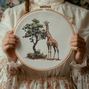Könnte beinhalten: Ein weißer Stickrahmen mit einem Kreuzstich-Motiv einer Giraffe, die neben einem Baum steht. Die Giraffe ist braun und gelb mit einem langen Hals und Flecken. Der Baum ist grün mit braunen Ästen.