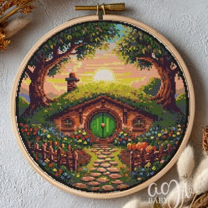 Può includere: Ricamo a punto croce di una scena di buca di hobbit, ambientato in un telaio di legno. L'opera presenta una porta verde, un sentiero di pietra e fiori colorati. Lo sfondo mostra un tramonto con alberi e un piccolo camino.