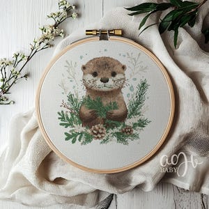 May include: A cross-stitch embroidery hoop with a cute otter holding a sprig of greenery. The otter is brown with a white chest and a white muzzle. The background is a light green with white snowflakes.