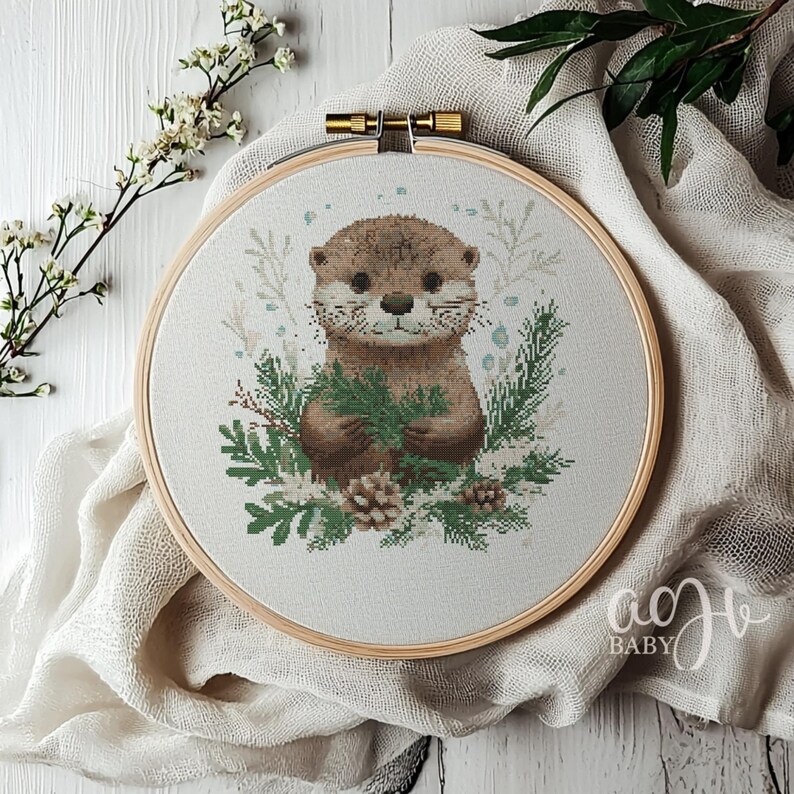 May include: A cross-stitch embroidery hoop with a cute otter holding a sprig of greenery. The otter is brown with a white chest and a white muzzle. The background is a light green with white snowflakes.