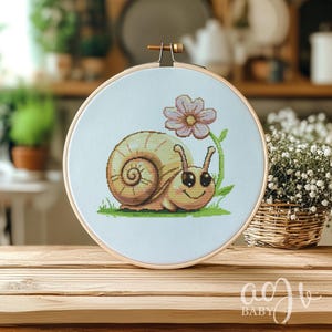 May include: A cross-stitch embroidery hoop with a light blue background featuring a cartoon snail with a brown shell and a pink flower. The snail has big, black eyes and a smiling mouth.