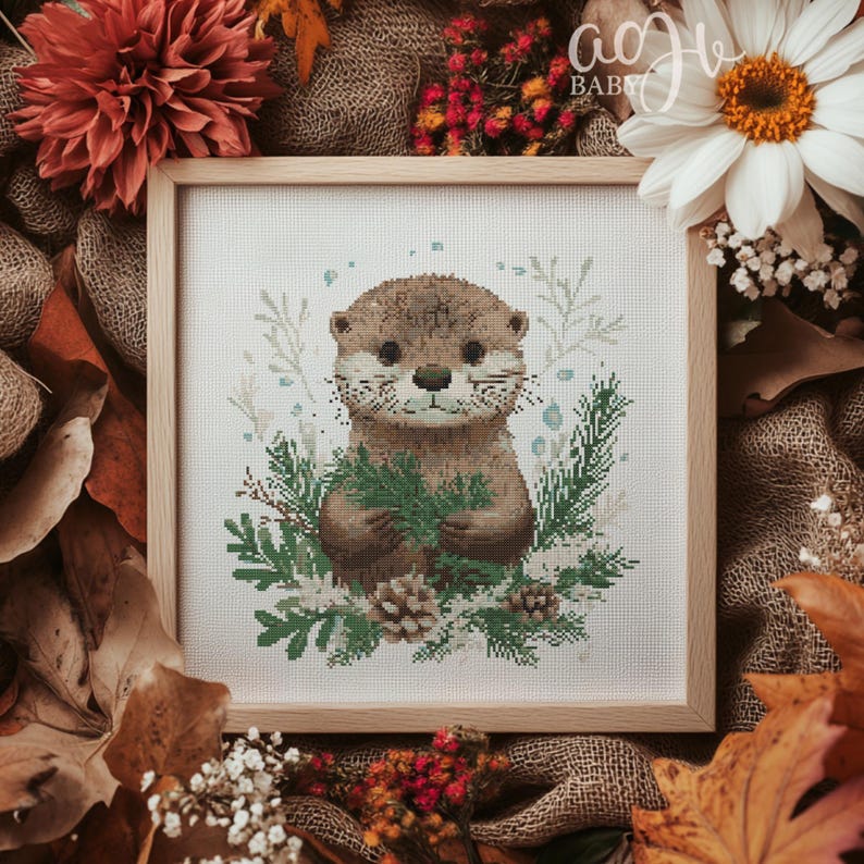 May include: A cross-stitch pattern featuring a cute otter holding a sprig of green leaves. The otter is surrounded by a floral design with white and green leaves. The background is white.