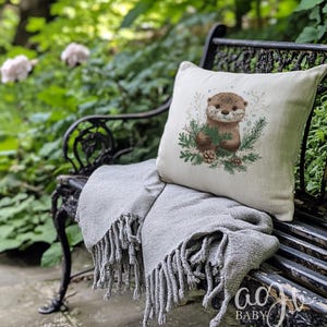 May include: A white throw pillow with a cute otter illustration holding a pine branch. The otter is surrounded by pine branches and pine cones. The pillow is sitting on a black metal bench with a gray throw blanket.