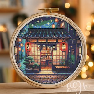 May include: A cross-stitch embroidery in a wooden hoop depicts a pixelated Japanese street scene at night. The scene includes a restaurant with red lanterns, a blue night sky, and a brick-paved street. The embroidery is framed in a round wooden hoop.