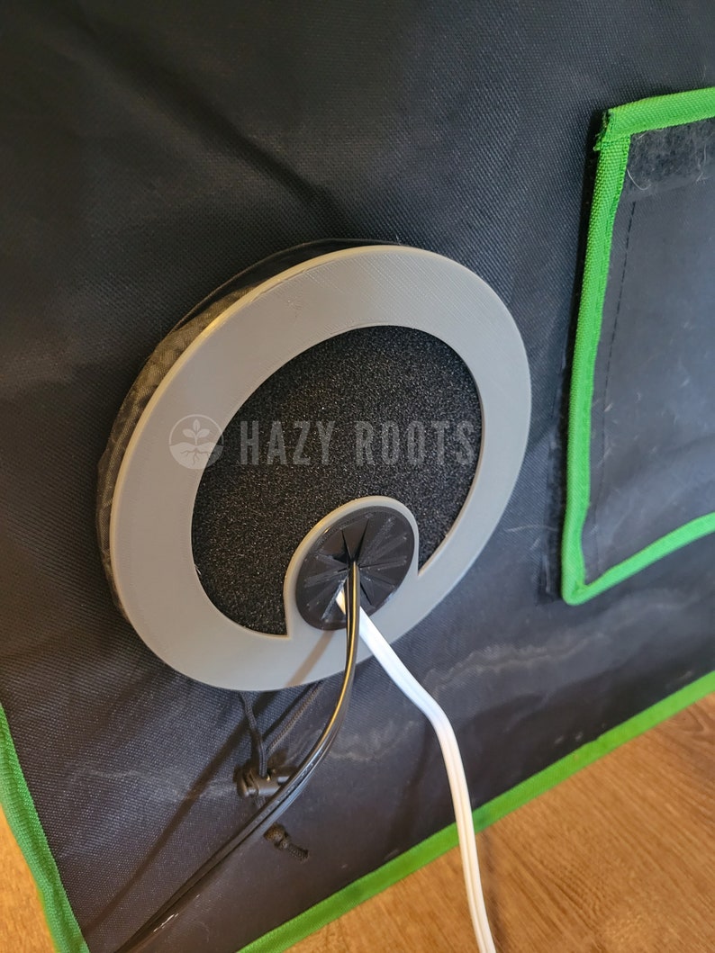 May include: A black and gray cable management grommet with a white cord and a black cord running through it. The grommet is attached to a black fabric surface with a green border.