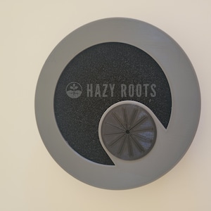 May include: Gray plastic disc with a black center and a cutout for a small, black, circular fan. The disc has the text "HAZY ROOTS" printed on it.