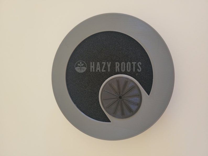 May include: Gray plastic disc with a black center and a cutout for a small, black, circular fan. The disc has the text "HAZY ROOTS" printed on it.