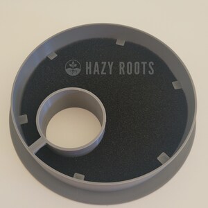 May include: Gray plastic plant pot saucer with a black foam insert and a central hole. The saucer has the text "HAZY ROOTS" printed on it.