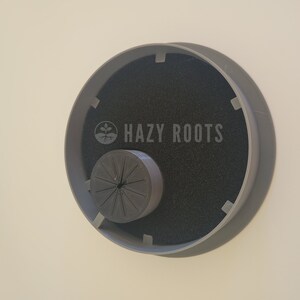 May include: A gray plastic plant pot with a black foam insert and a gray plastic ring with the text "HAZY ROOTS" printed on it.