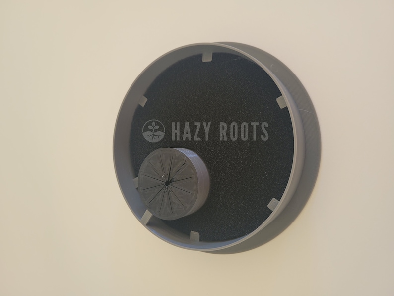 May include: A gray plastic plant pot with a black foam insert and a gray plastic ring with the text "HAZY ROOTS" printed on it.