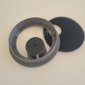 May include: A gray plastic ring with a black foam filter and a black plastic insert with a starburst design. The ring has four slots on the inside. The text "HAZY ROOTS" is visible on the ring.