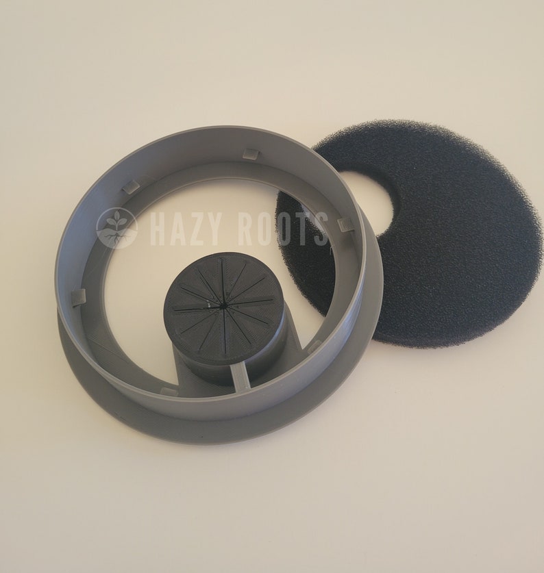 May include: A gray plastic ring with a black foam filter and a black plastic insert with a starburst design. The ring has four slots on the inside. The text "HAZY ROOTS" is visible on the ring.