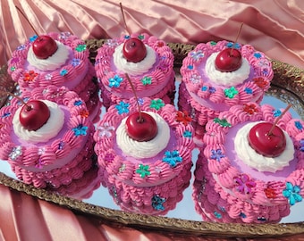 Puede incluir: Seis mini pasteles rosas y blancos con cerezas rojas en la parte superior. Cada pastel está decorado con chispas y glaseado de colores.