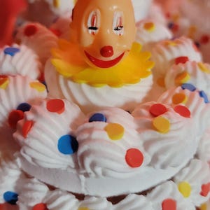 May include: A whimsical, multi-tiered dessert featuring a clown figurine. The dessert is decorated with white frosting swirls and colorful sprinkles. The clown has a red hat, yellow collar, and a smiling face. The overall theme is festive and playful.