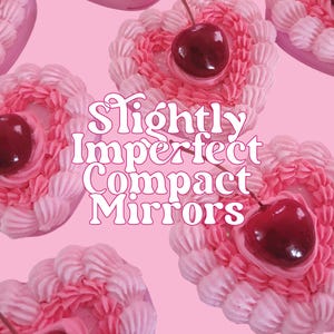 Könnte beinhalten: Eine Sammlung von herzförmigen Kompakt-Spiegeln in Rosa und Weiß mit einer Kirsche oben drauf. Der Text "Slightly Imperfect Compact Mirrors" ist in rosa und weißen Buchstaben geschrieben.