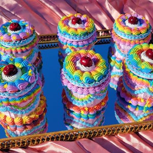 May include: A collection of colorful, layered, rainbow-frosted cakes with a cherry on top, presented on a mirrored tray with a gold-colored frame. The cakes are arranged on a pink satin surface.