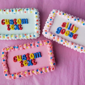 以下が含まれることがあります： 白、ピンク、白のフロスティングを施した3つの長方形のケーキ。各ケーキには、カラフルなドットが付いた白いフロスティングのボーダーがあります。1つのケーキにはカラフルな文字で「custom text」と書かれており、別のケーキには「silly goose」と書かれています。