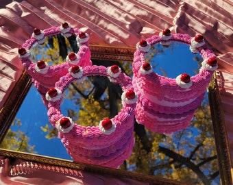 Puede incluir: Tres espejos decorativos en forma de pastel de varios pisos, de color rosa y blanco, con cerezas rojas en la parte superior. Los espejos se reflejan en un espejo con marco dorado.