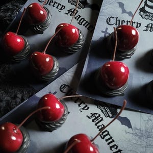 Op de afbeelding: Close-up van verschillende kersmagneten met een glanzende rode afwerking en bruine stelen. De magneten zijn bevestigd aan grijze bases en worden gepresenteerd op kaarten met de tekst "Cherry Magnets". De achtergrond is donker en getextureerd.