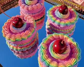 Puede incluir: Cuatro pequeños pasteles rosas en capas con glaseado arcoíris y una sola cereza roja encima. Los pasteles están dispuestos sobre una superficie espejada, que refleja el cielo azul y los propios pasteles. El glaseado está decorado.