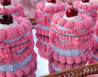 Puede incluir: Cuatro pasteles de capas rosas y blancas con cerezas en la parte superior, exhibidos en una bandeja dorada.