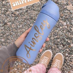 Peut inclure: Une gourde bleu clair avec le nom "Ainsley" en lettres dor&eacute;es. La gourde a un couvercle bleu. Le texte "FREE SHIPPING" est dans un rectangle beige. La gourde est tenue par une personne.