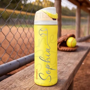 K&ouml;nnte beinhalten: Eine gelbe Owala-Trinkflasche mit einem Softball-Motiv und dem Namen "Sophia" in silberner Schrift. Die Flasche hat einen wei&szlig;en und gelben Deckel und steht auf einer Holzbank. Ein Softball und ein Handschuh sind im Hintergrund.