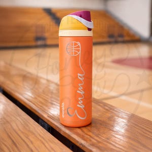 K&ouml;nnte beinhalten: Eine orangefarbene, isolierte Wasserflasche mit einem Basketball-Motiv und dem Namen "Emma" in Schreibschrift. Der Deckel ist gelb und lila. Die Flasche steht auf einer Holzbank in einer Turnhalle.