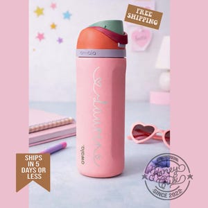 K&ouml;nnte beinhalten: Eine rosa Owala-Trinkflasche mit einem mehrfarbigen Deckel. Die Flasche tr&auml;gt den Namen "Laura" in Schreibschrift. Die Flasche ist zylindrisch und hat eine strukturierte Oberfl&auml;che. Der Deckel ist orange, gr&uuml;n und lila. Die Flasche steht auf einem Schreibtisch mit einem Notizbuch und einer Sonnenbrille.