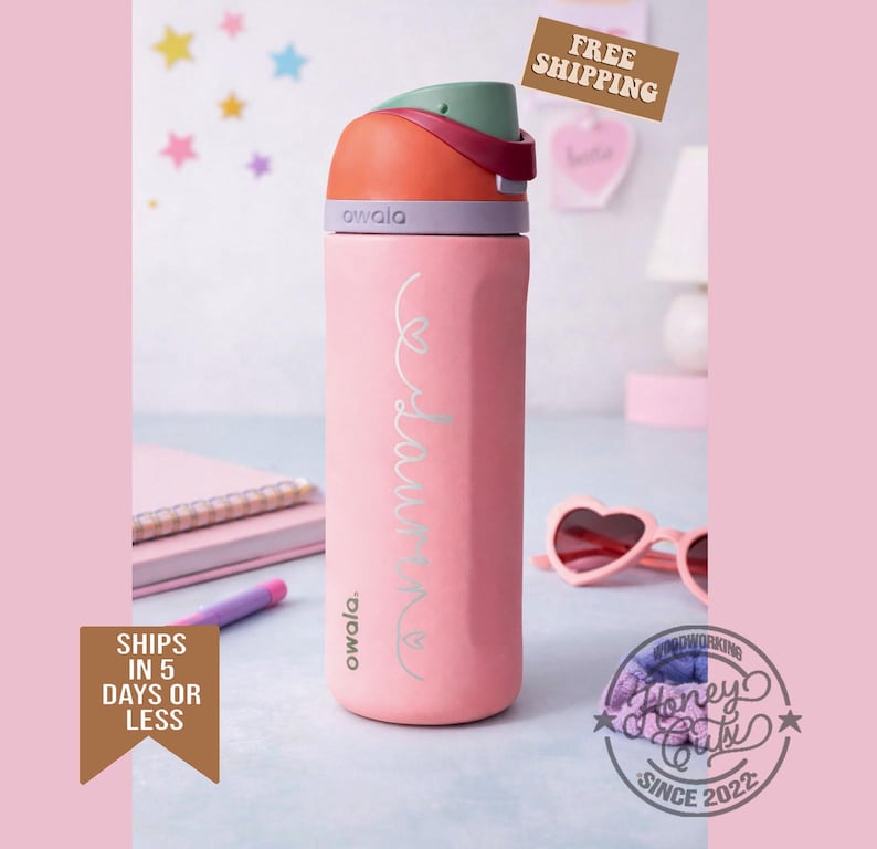 K&ouml;nnte beinhalten: Eine rosa Owala-Trinkflasche mit einem mehrfarbigen Deckel. Die Flasche tr&auml;gt den Namen "Laura" in Schreibschrift. Die Flasche ist zylindrisch und hat eine strukturierte Oberfl&auml;che. Der Deckel ist orange, gr&uuml;n und lila. Die Flasche steht auf einem Schreibtisch mit einem Notizbuch und einer Sonnenbrille.