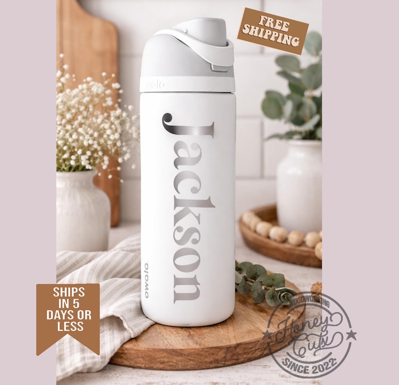 K&ouml;nnte beinhalten: Eine wei&szlig;e Owala-Trinkflasche mit grauem Deckel und dem Namen "Jackson" in silbernen Buchstaben. Die Flasche steht auf einem Holzschneidebrett, mit einer Vase mit wei&szlig;en Blumen und Gr&uuml;nzeug im Hintergrund.