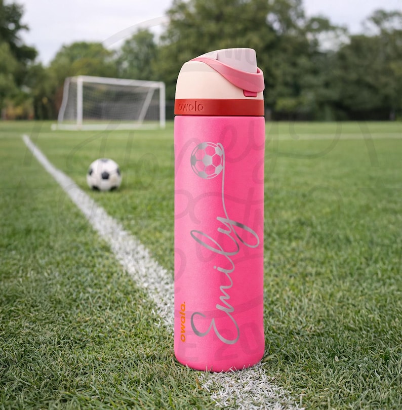 K&ouml;nnte beinhalten: Eine pinkfarbene Owala-Trinkflasche mit einem Fu&szlig;ball-Motiv und dem Namen "Emily" in silberner Schrift. Die Flasche hat einen wei&szlig;en und rosa Deckel und ein rotes Band. Ein Fu&szlig;ball und ein Tor befinden sich im Hintergrund auf einem gr&uuml;nen Feld.