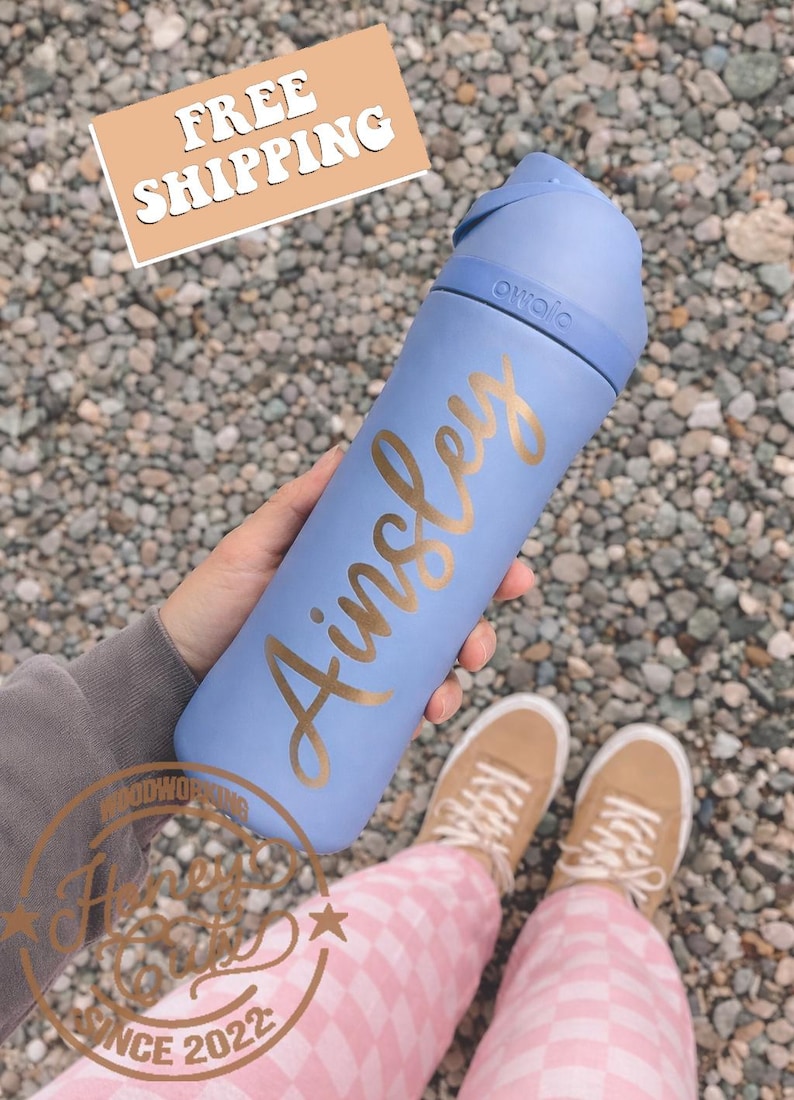 Peut inclure: Une gourde bleu clair avec le nom "Ainsley" en lettres dor&eacute;es. La gourde a un couvercle bleu. Le texte "FREE SHIPPING" est dans un rectangle beige. La gourde est tenue par une personne.