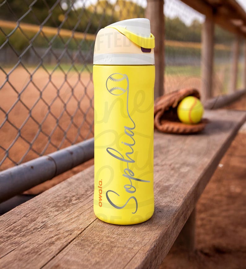 K&ouml;nnte beinhalten: Eine gelbe Owala-Trinkflasche mit einem Softball-Motiv und dem Namen "Sophia" in silberner Schrift. Die Flasche hat einen wei&szlig;en und gelben Deckel und steht auf einer Holzbank. Ein Softball und ein Handschuh sind im Hintergrund.
