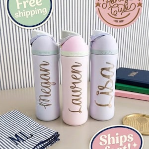 Op de afbeelding: Drie pastelkleurige Owala waterflessen met de namen Megan, Lauren en Lisa in gouden letters. De flessen staan op een tafel met een gestreepte doek en decoratieve stickers. De flessen hebben een klapdeksel.