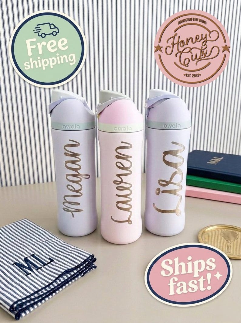 Op de afbeelding: Drie pastelkleurige Owala waterflessen met de namen Megan, Lauren en Lisa in gouden letters. De flessen staan op een tafel met een gestreepte doek en decoratieve stickers. De flessen hebben een klapdeksel.