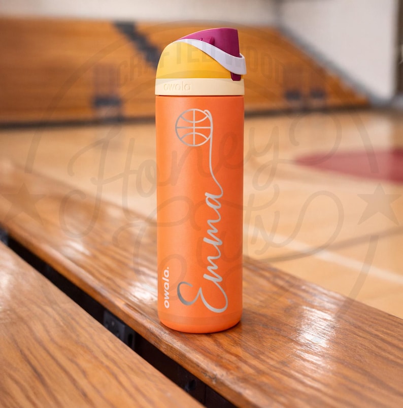 K&ouml;nnte beinhalten: Eine orangefarbene, isolierte Wasserflasche mit einem Basketball-Motiv und dem Namen "Emma" in Schreibschrift. Der Deckel ist gelb und lila. Die Flasche steht auf einer Holzbank in einer Turnhalle.