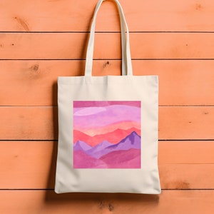 May include: A white canvas tote bag featuring a pink and purple mountain landscape design. The mountains are layered and have a watercolour effect.