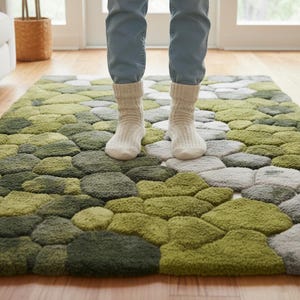 May include: A green and gray area rug with a pebble design. The rug is made of a soft, textured material and features various shades of green and gray. A person is standing on the rug, wearing white socks and blue jeans. The rug is on a wooden floor.